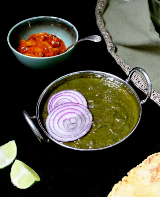A bowl with sarson da saag and a red lime pickle on the side on a black table with a green napkin