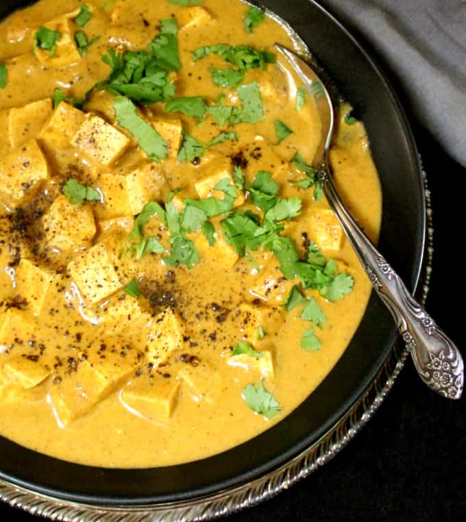 A close up top shot of a black bowl filled with a creamy yellow Tofu Paneer Kalimirch, a dish of tofu cubes in a spicy black pepper sauce. 
