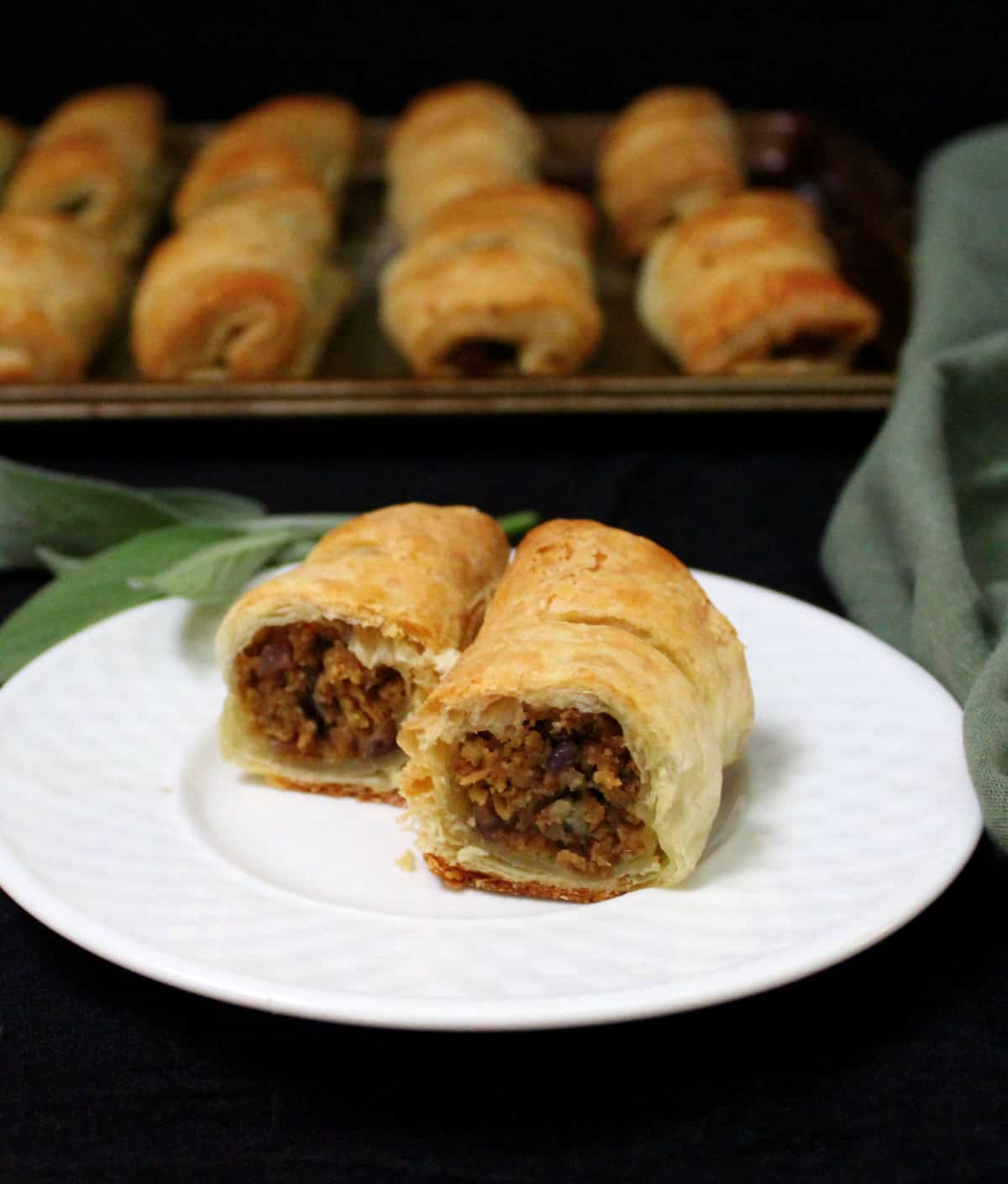 A sliced sausage roll on a white plate in the foreground with more sausage rolls on a baking sheet in the back against a black background
