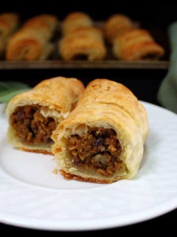 Golden, flaky vegan sausage rolls on a white plate stuffed with apple sage sausage and with more rolls in the background on a baking sheet