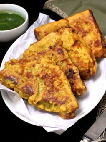 Four golden brown bread pakoras lined up on a white plate with green chutney in the background and a green napkin