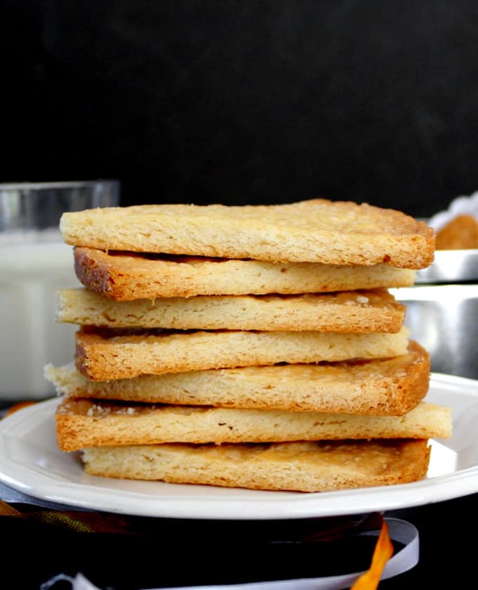 Photo of six shortbread cookies stacked on top of a white plate with a glass of milk