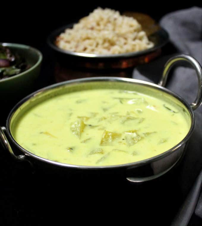 Front shot of a steel bowl of creamy green tomato stew with mushroom stir-fry and brown jeera rice on a black background