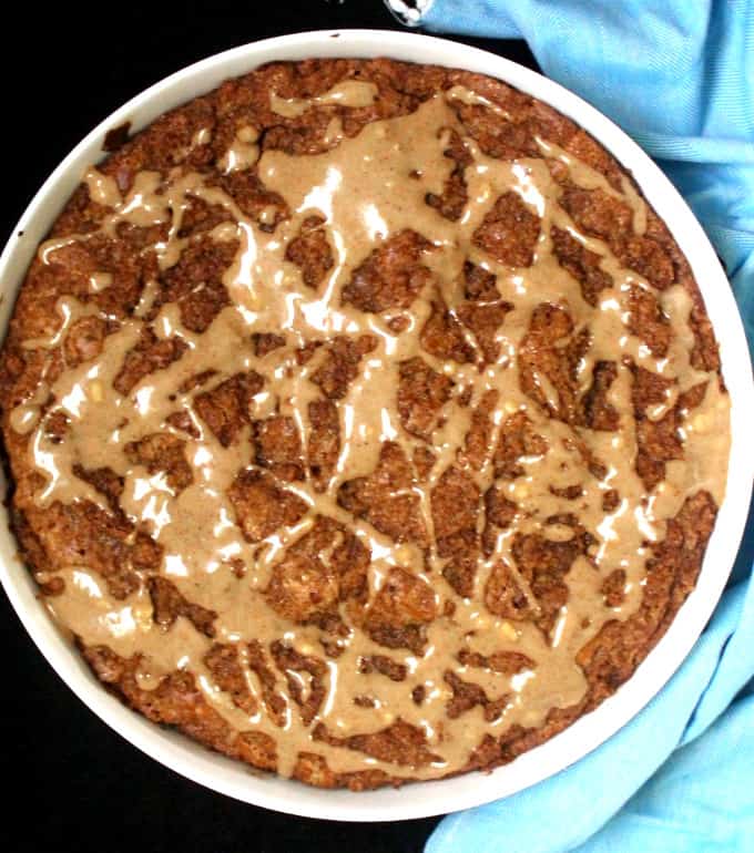 A large vegan cinnamon cake with vegan streusel and vanilla cashew glaze in a white round baking pan with a blue napkin beside it
