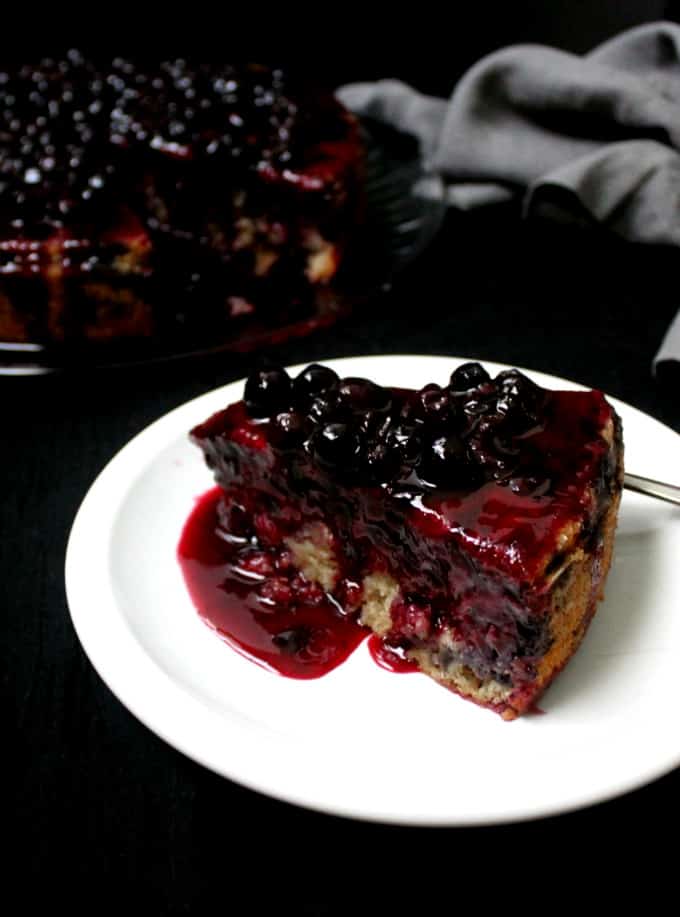 A slice of vegan lemon blueberry cake on a white plate with blueberries on top and the topping dripping down the sides and the full cake in the background