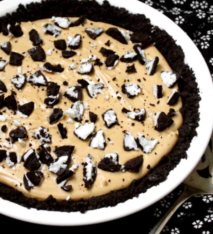A front partial shot of a decadent peanut butter oreo pie in a white plate with cookies crumbled on top