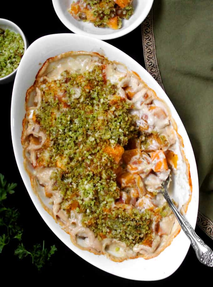 A top shot of an oval white baking dish with a vegan butternut squash gratin and a silver spoon with a green napkin and parsley to the side.
