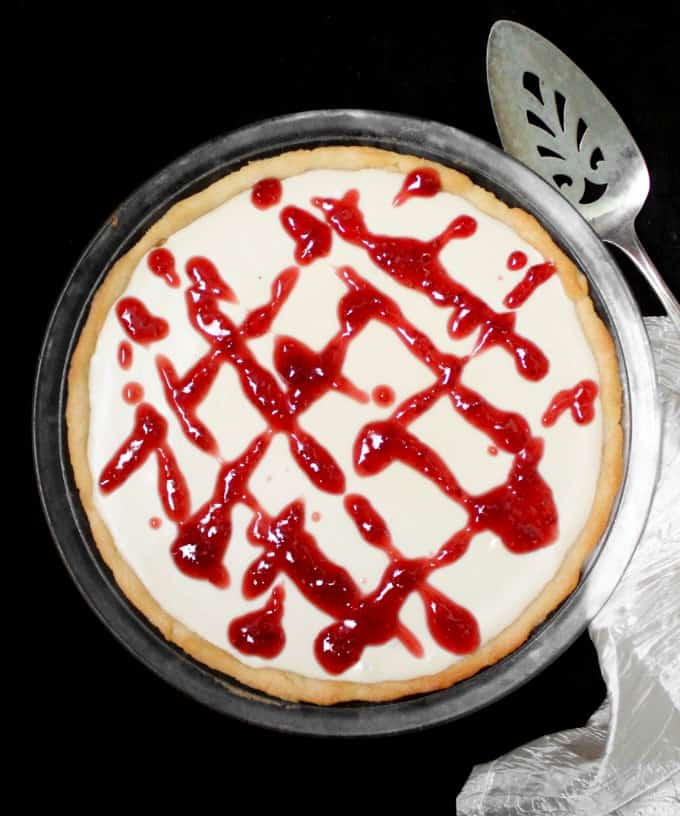 A top shot of a vegan white chocolate pie with an almond shortbread crust and a raspberry drizzle with a silver server next to it