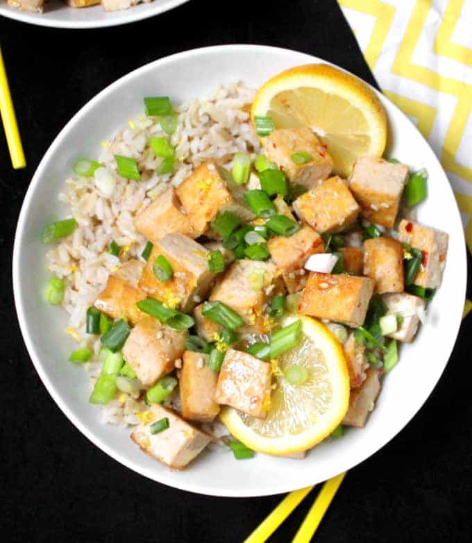 A bowl of sticky glazed lemon tofu with brown rice and scallions and slices of lemon in a white bowl with yellow chopsticks.