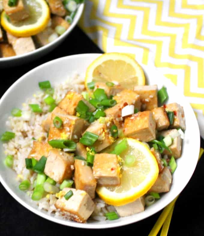 A white bowl with brown rice and glazed lemon tofu with scallions sprinkled on top and yellow chopsticks and napkin beside it.