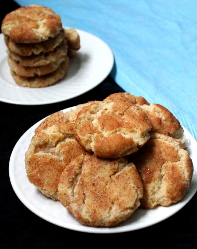 Several vegan snickerdoodle cookies piled on two white plates with a blue napkin next to them