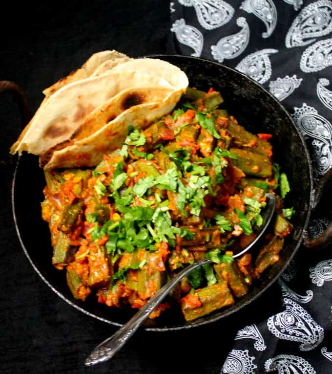 Bhindi Masala is an Indian style vegetarian dish of okra with a tomato onion sauce. Here you see it in a black Indian wok with a silver spoon and a black and white napkin