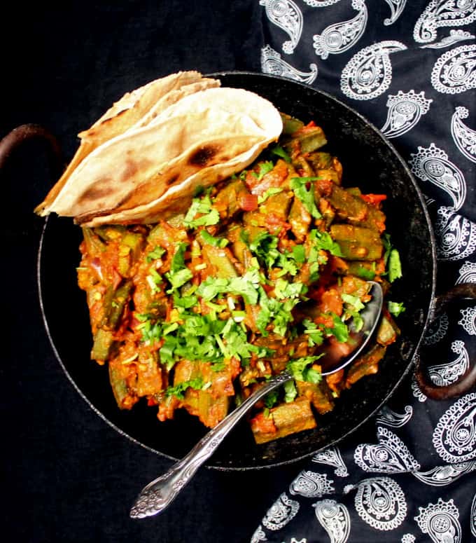Top shot of bhindi masala with rotis tucked in the black bowl and a spoon