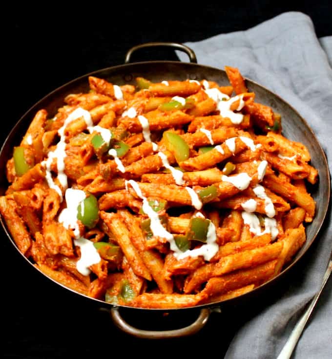 A front shot of Indian style masala pasta with green bell peppers and cashew cream in a big copper pan with a gray napkin on a black background.