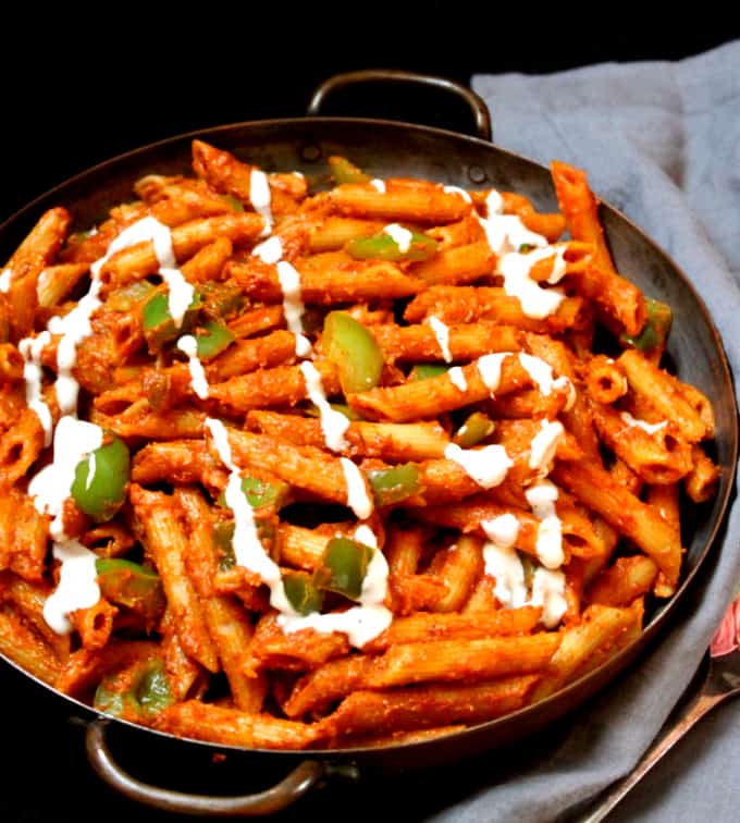 Close up photo of a fiery red masala pasta made in Indian street food style with a drizzle of cashew cream and bell peppers in a copper pan with a gray napkin on a black background.