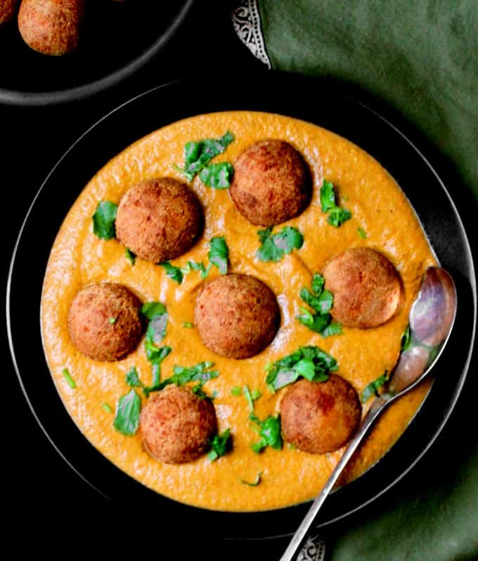 Vegan malai kofta in a black bowl with a silver spoon and fried kofta meatballs to the side against a green napkin.