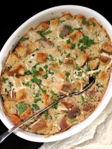 An overhead shot of a white, oval casserole dish with spinach, sausage and cheese casserole and a serving spoon on black background