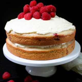 A front shot of a vegan white chocolate raspberry cake on a cake stand with fresh raspberries piled on top