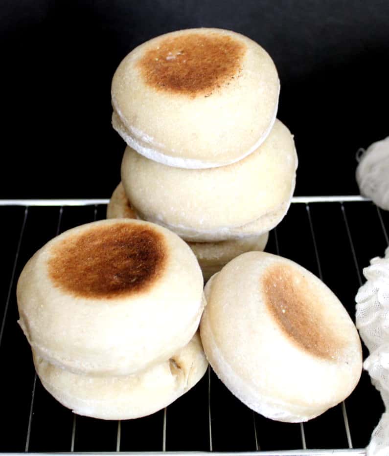 Stacks of Sourdough English Muffins perfectly baked cooling on a rack on a black background next to cheesecloth.