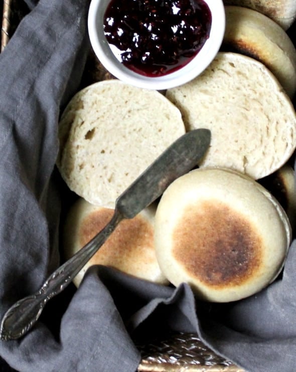 Close up of sourdough English Muffins in a wicker basket with a gray napkin and raspberry jam and a butter knife.