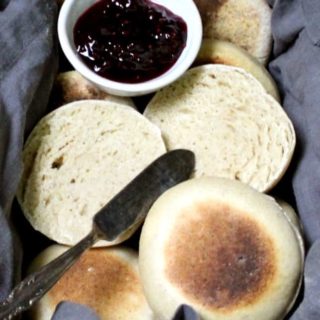 A close up of sourdough English muffins in a wicker basket with gray napkin, raspberry jam and a butter knife