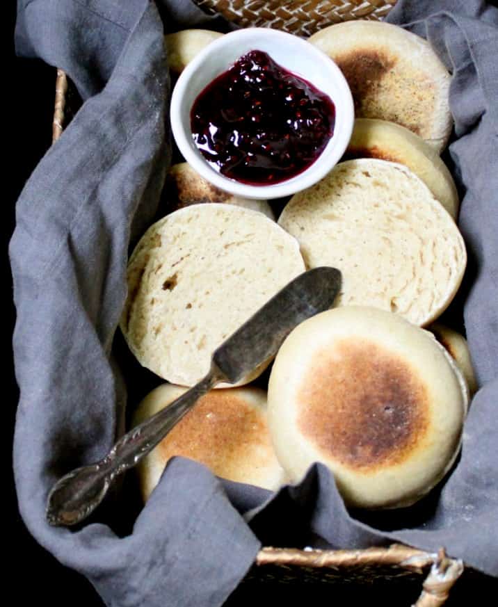 Close up of sourdough English Muffins in a wicker basket with a gray napkin and raspberry jam and a butter knife.
