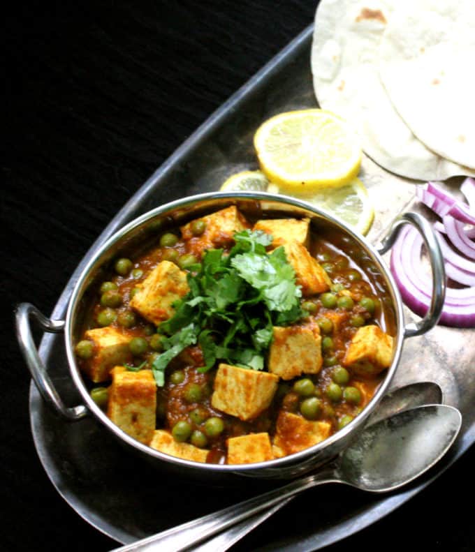 A front shot of matar paneer with tofu in a steel karahi bowl with lemon, roti, onions and two silver spoons.
