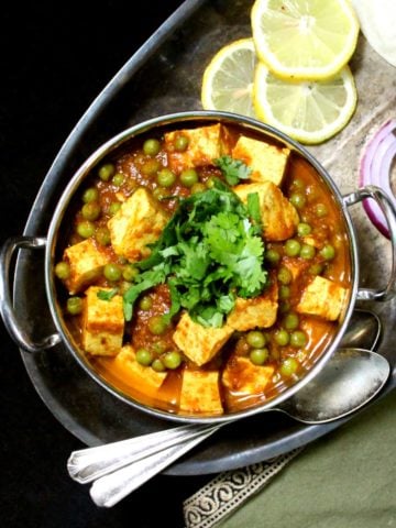 A kadhai serving bowl with matar paneer with tofu cubes in a tomato onion gravy sauce, with onions, lemons and roti