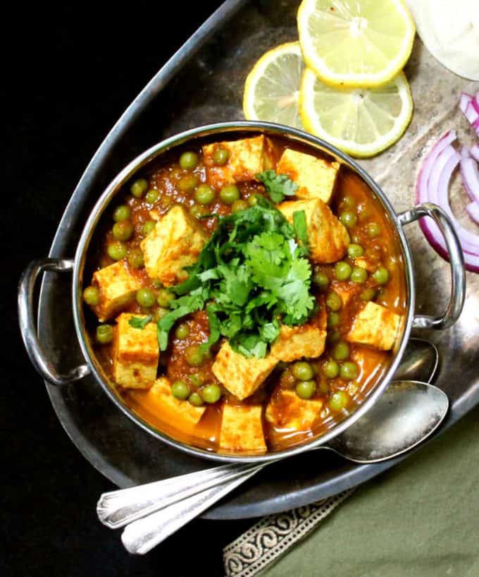 A kadhai bowl with mater paneer with cubes of tofu and green peas on a silver tray with wedges of lemon, roti and slices of onion.