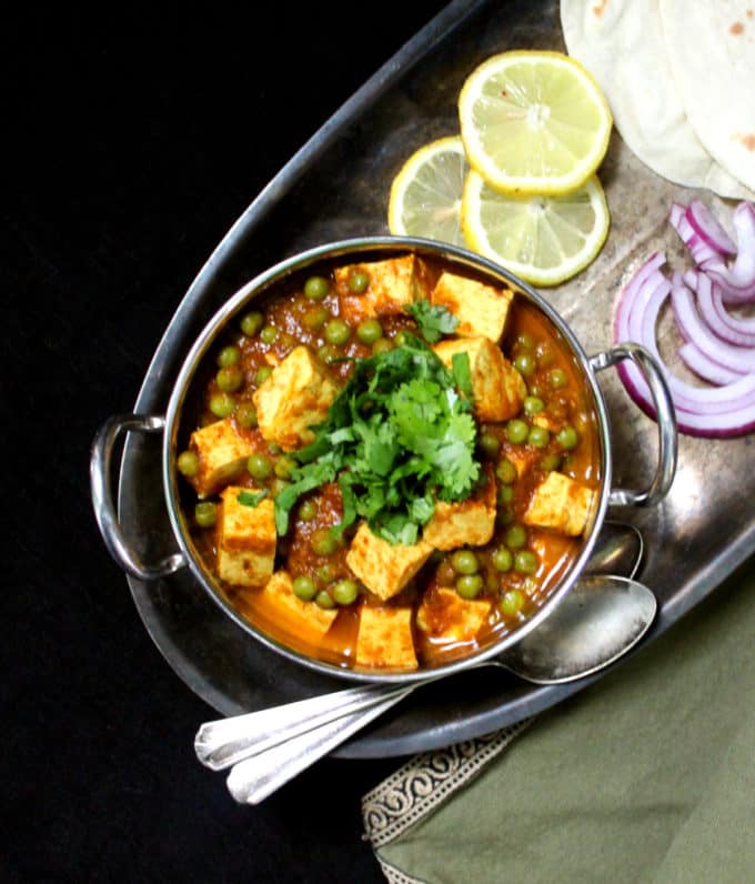 Overhead shot of matar paneer with lemon wedges, slices of onion, rotis and two spoons.