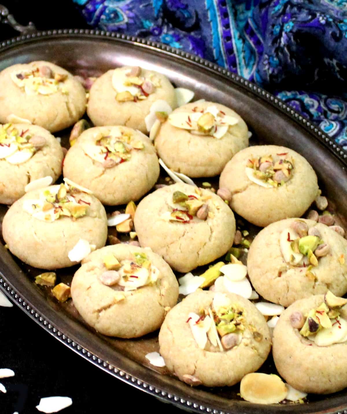 A close up shot of several vegan nankhatai biscuits on a silver plate with pistachios, almonds, saffron and other garnishes.