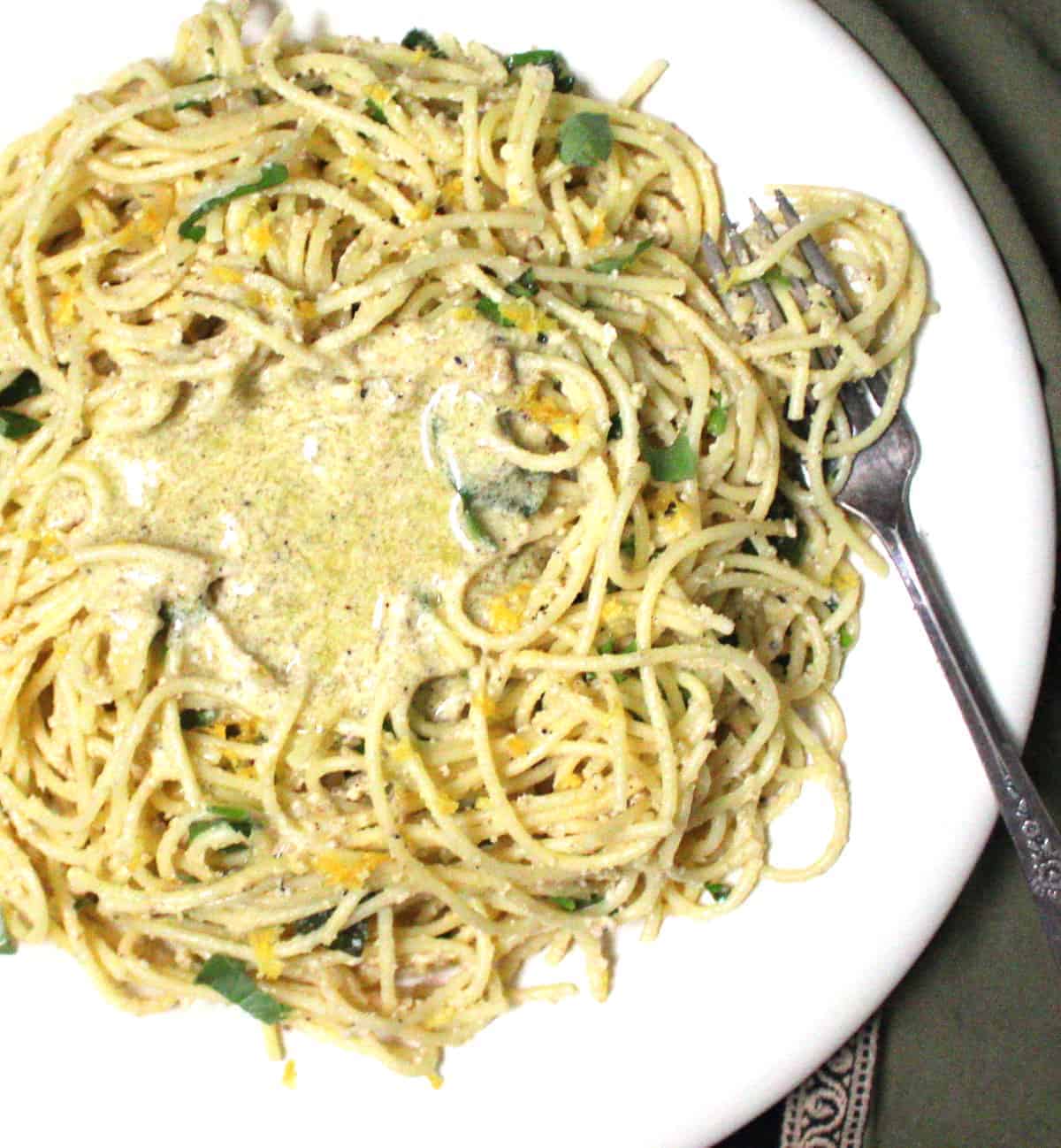 An overhead closeup of vegan lemon spaghetti pasta with shards of lemon zest, basil, and creamy lemon sauce on a white plate with a silver fork and green and gold napkin.