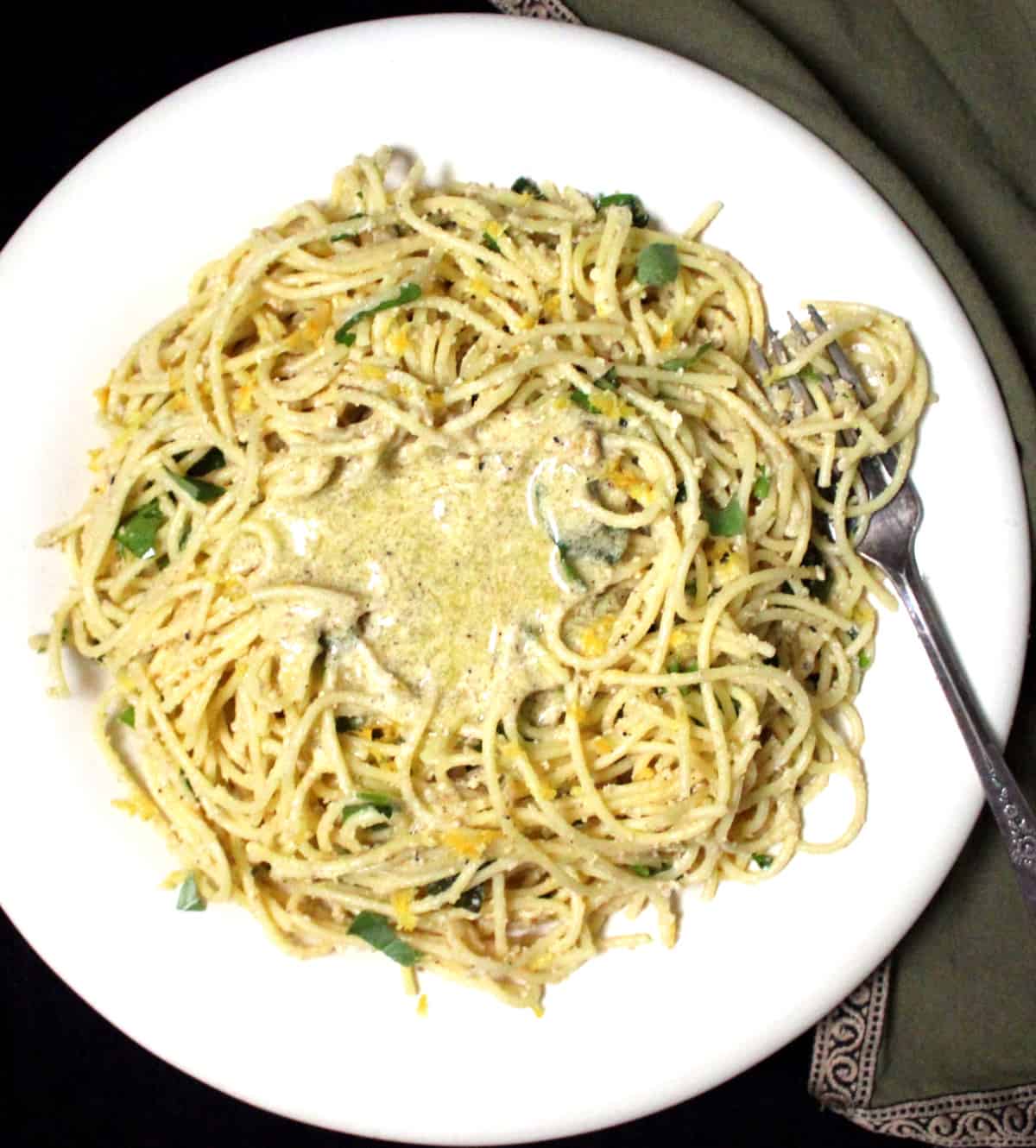 An overhead shot of vegan lemon spaghetti pasta with shards of lemon zest, basil, and creamy lemon sauce on a white plate with a silver fork and green and gold napkin.