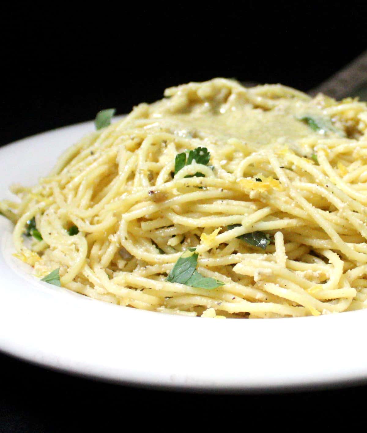 A front close up of vegan lemon spaghetti with parsley, basil, vegan parmesan and lemon zest on a white plate with a green napkin.