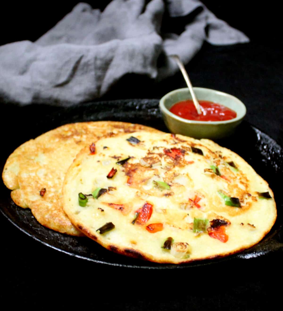 Front shot of two moonglets or mung bean omelets studded with veggies like bell peppers, onions and scallions on a cast iron griddle.