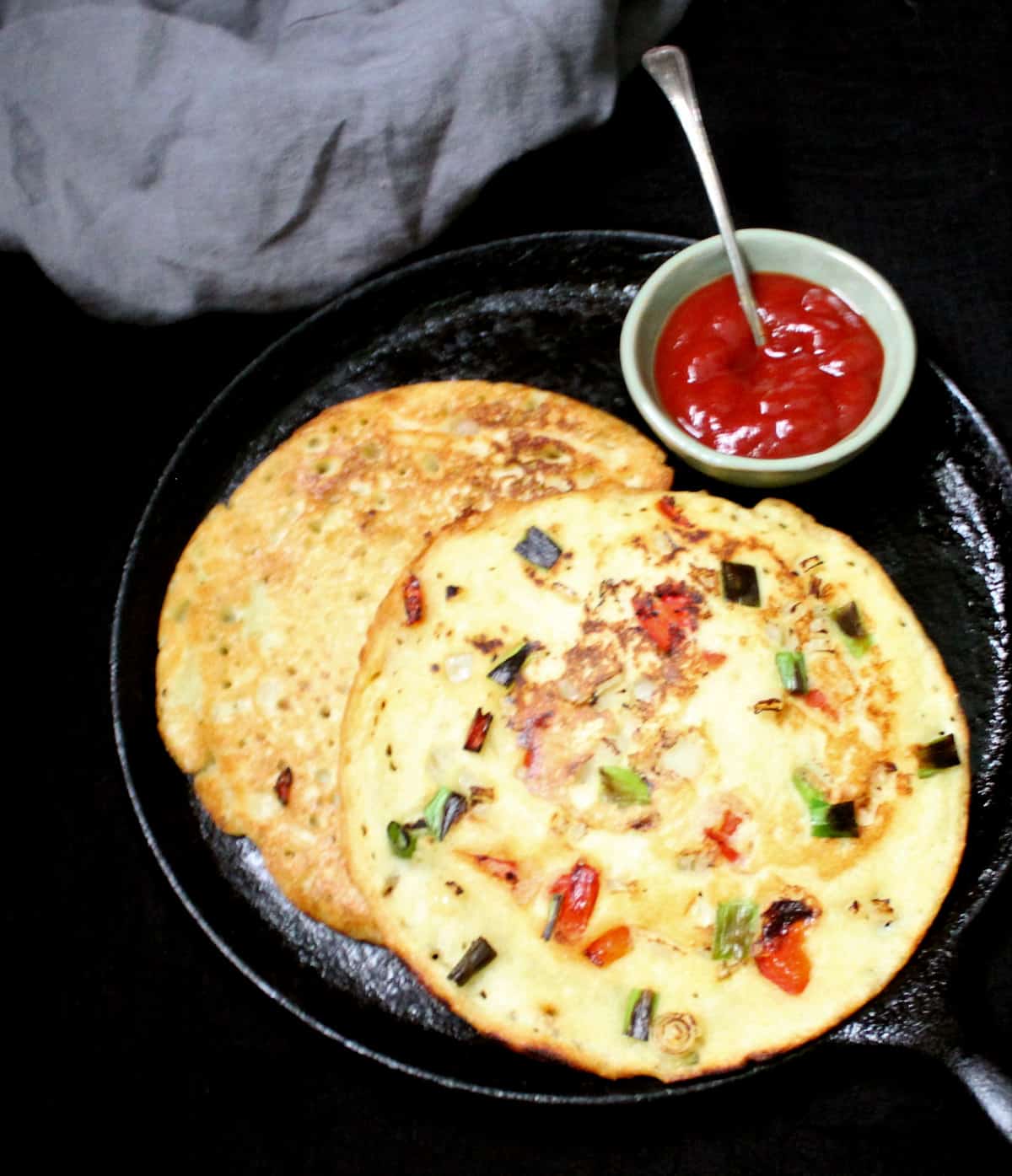 Overhead shot of Indian style moonglets or munglets with bits of vegetables on a cast iron griddle with ketchup and a gray napkin in the background.