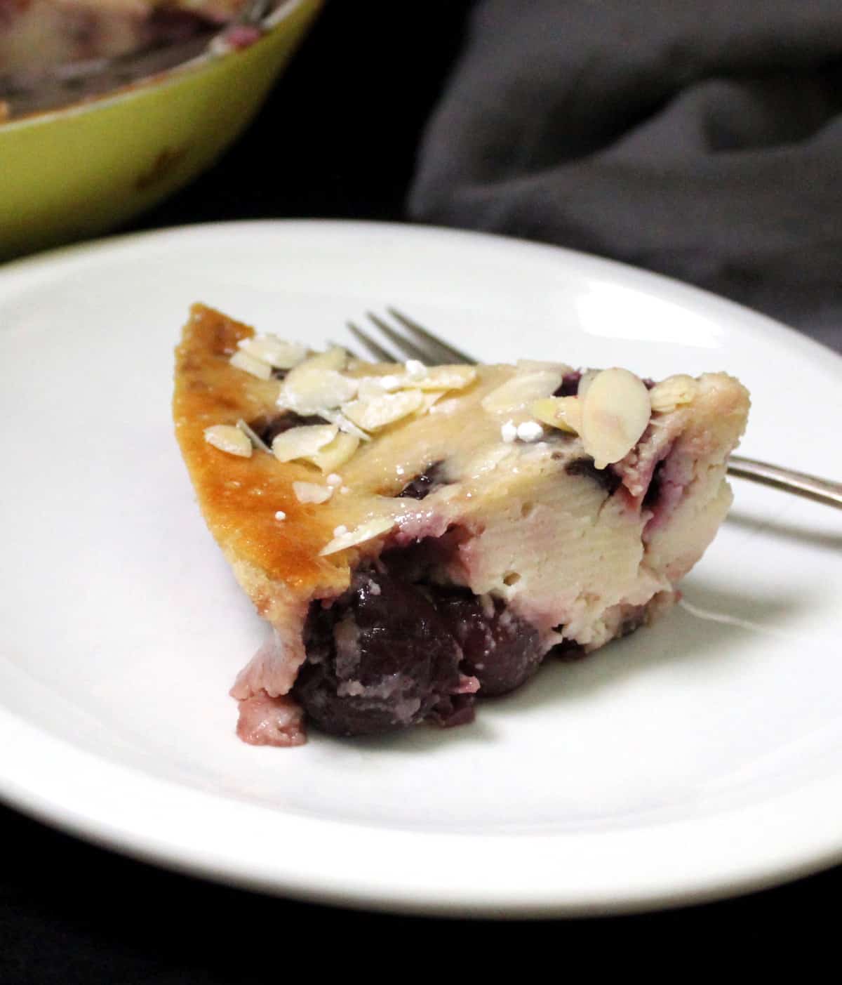 Photo of a slice of vegan cherry clafoutis on a white plate with a fork.