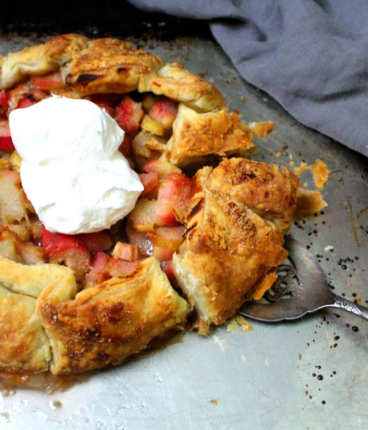 Close of vegan rhubarb galette with a slice held up by a silver pie server and vegan whipped cream.