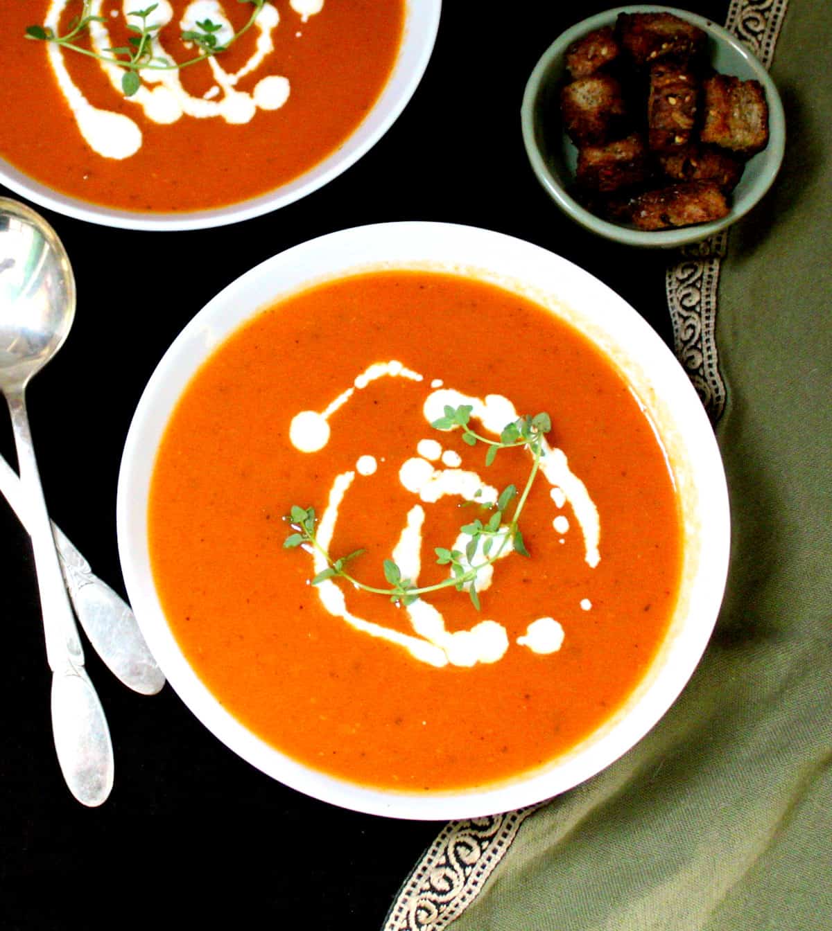 An overhead shot of two bowls of dairyfree vegan tomato soup with thyme, cashew cream, croutons and silver spoons.