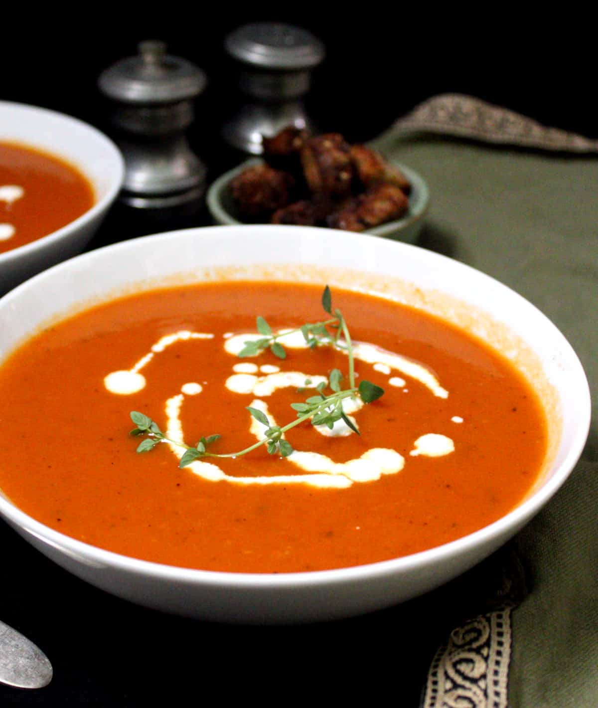 A front partial shot of a bowl of bright red tomato soup with sprigs of thyme, cashew cream and croutons on the side, and salt and pepper shakers and a green napkin beside.