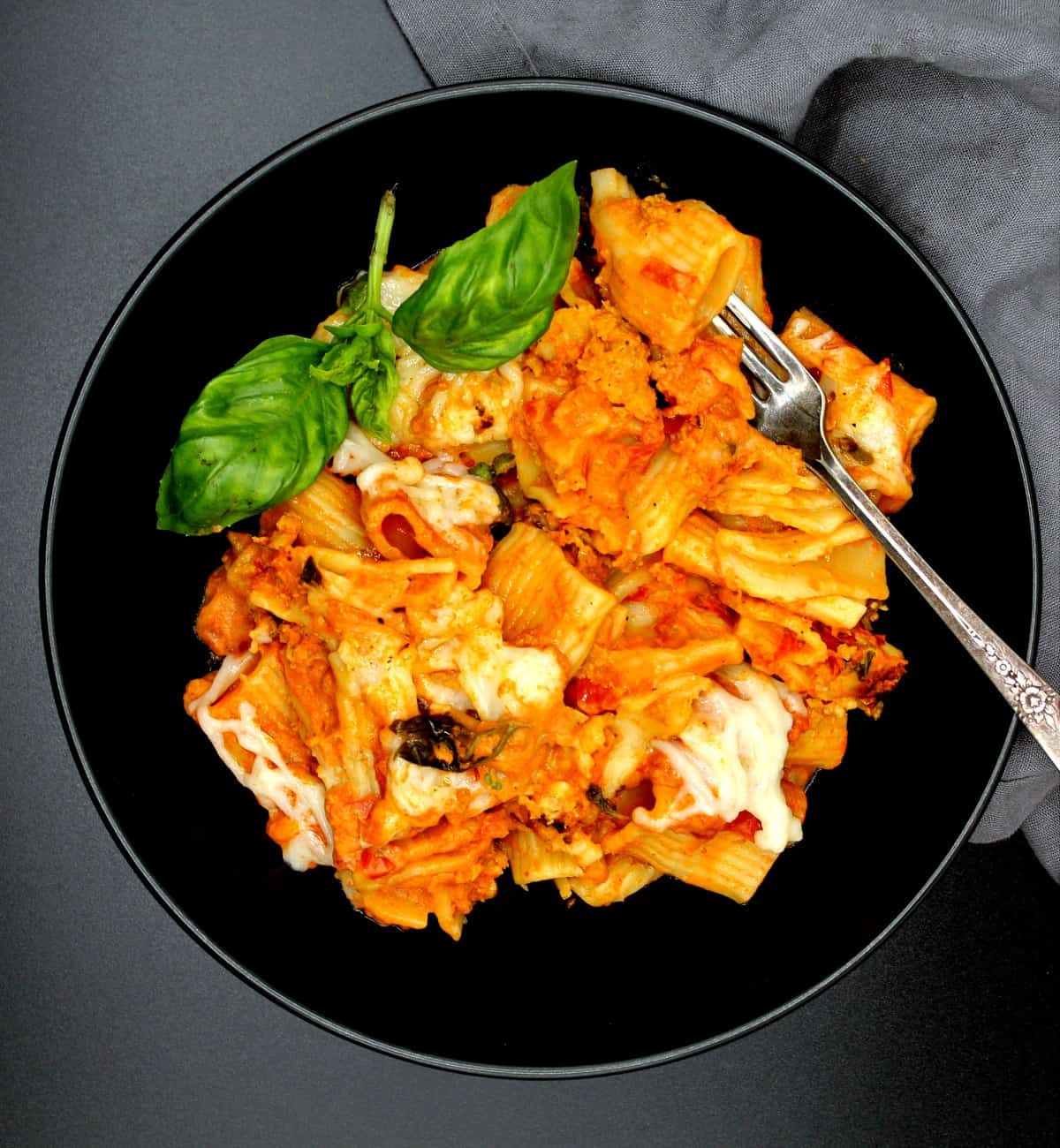 An overhead shot of a black bowl with a serving of creamy, cheesy vegan pasta bake with rigatoni, shreds of vegan mozzarella cheese and a gray napkin.