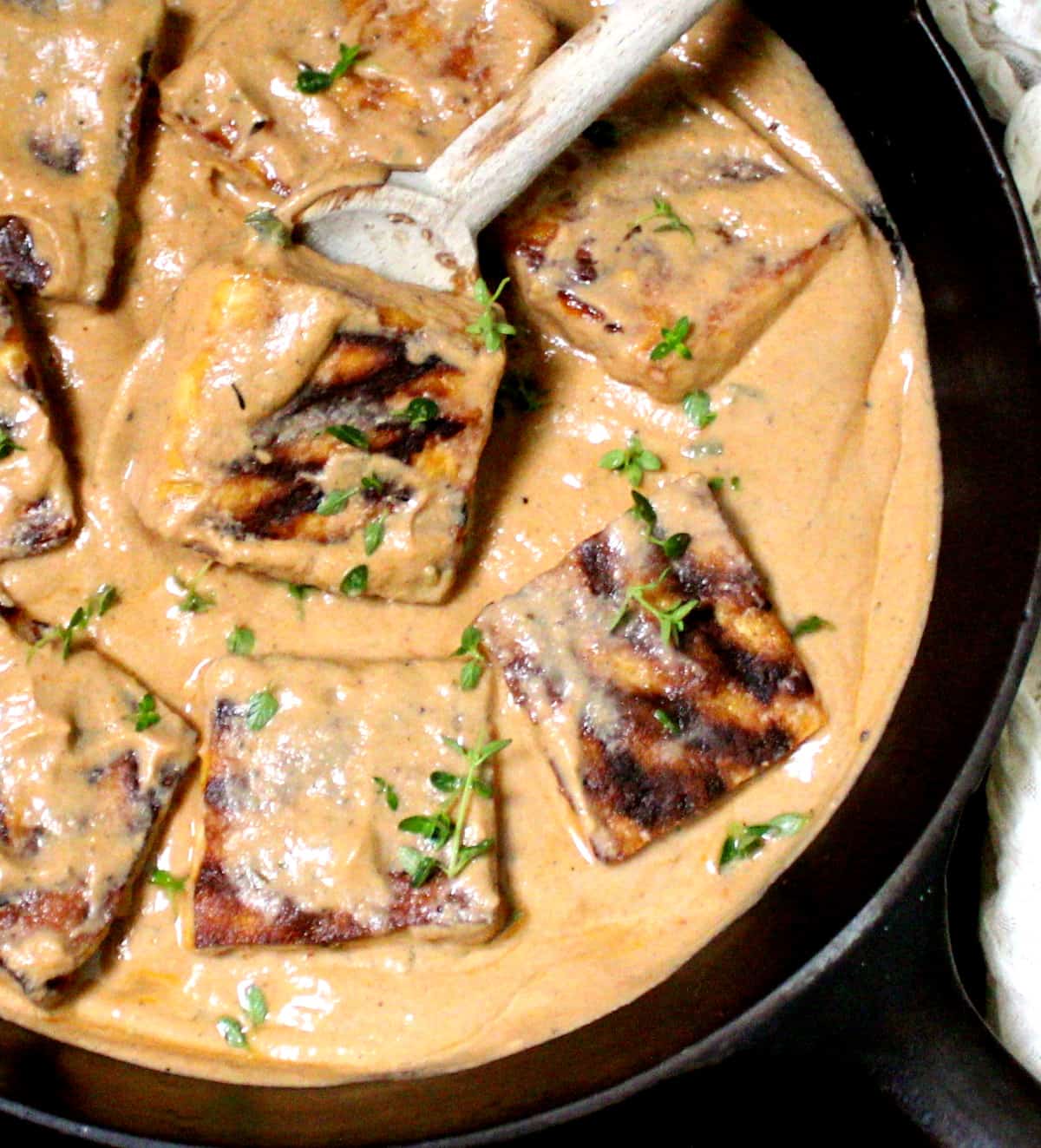 Overhead closeup of grilled tofu steaks in a creamy vegan Cajun sauce in a cast iron skillet with a wooden ladle and cheesecloth