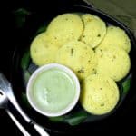 Yellow rava idlis made with vegan yogurt on a black plate with cilantro coconut chutney and a knife and fork