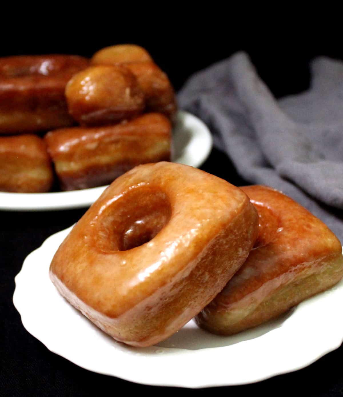 Front shot of two square donuts with a stack of donuts on a white plate and a gray napkin.