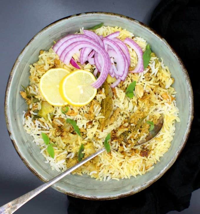 Overhead shot of a bowl of keema biryani, an Indian rice dish with ground meatless meat and spices, in a glazed bowl with a spoon, lemons and sliced onions