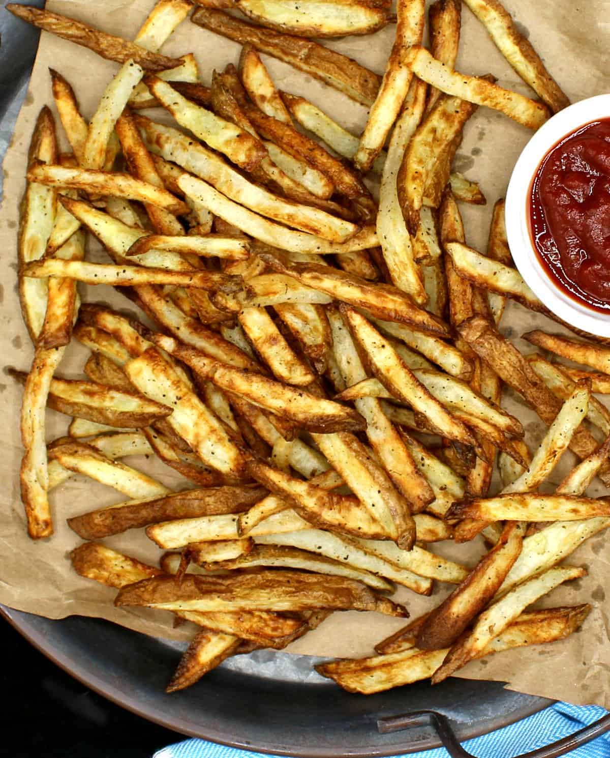 A closeup top photo of air fryer French fries, golden and crispy, on brown paper. These are made with very little oil or no oil. Next to the fries is a white ceramic bowl with tomato ketchup.