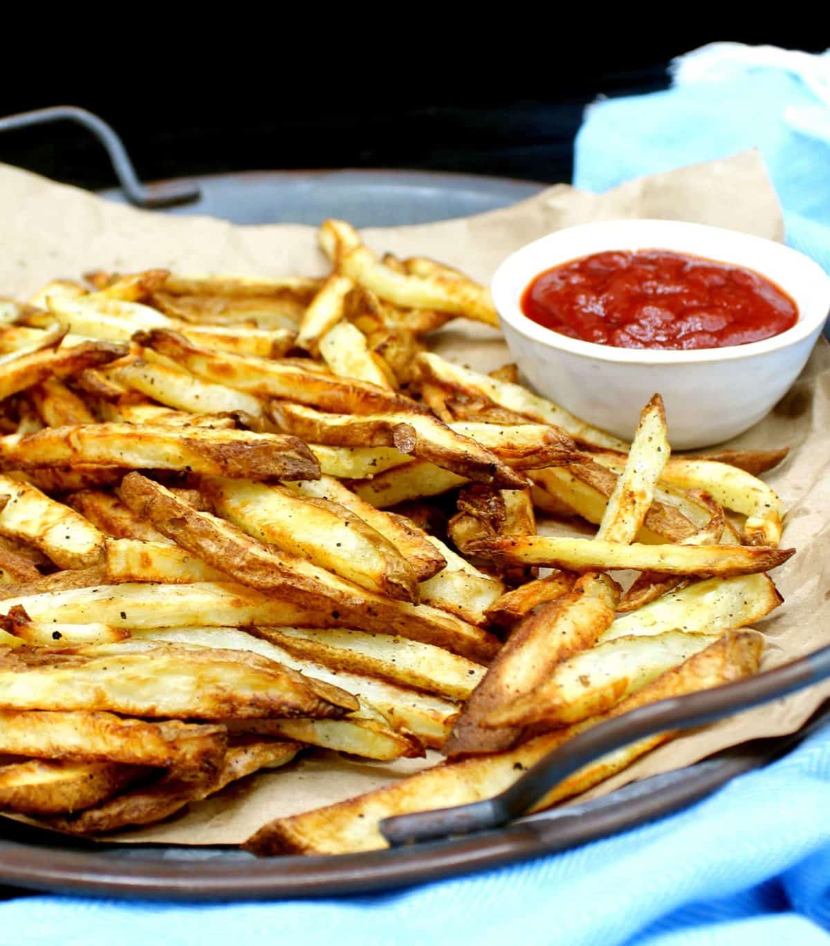 A front closeup of air fryer French fries, golden and crispy, on brown paper. These are made with very little oil or no oil. Next to the fries is a white ceramic bowl with tomato ketchup.