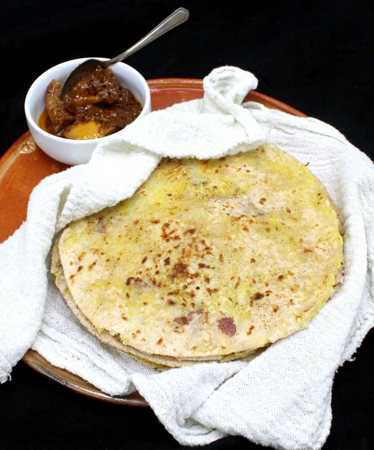A front shot of a stack of freshly baked aloo parathas nestled in a white flour sack towel on an earthenware plate with a small white ceramic bowl of Indian lime pickle on the side, all on a black background.