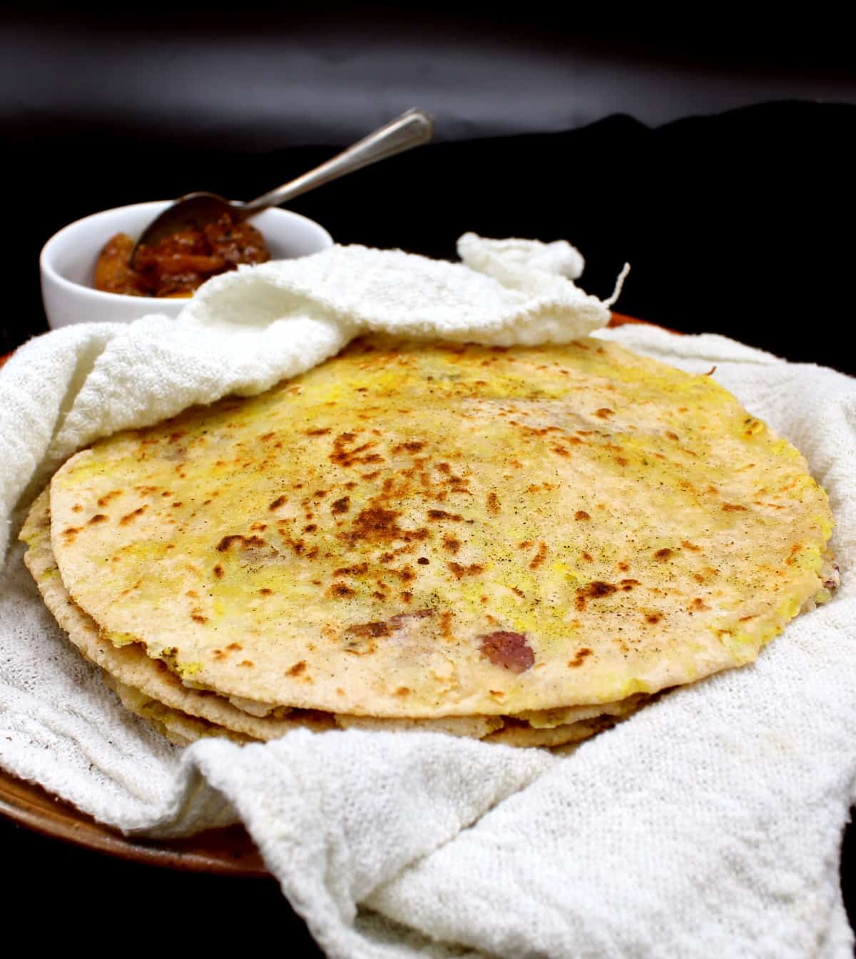 A front shot of a stack of freshly baked aloo parathas nestled in a white flour sack towel on an earthenware plate with a small white ceramic bowl of Indian lime pickle on the side, all on a black background.
