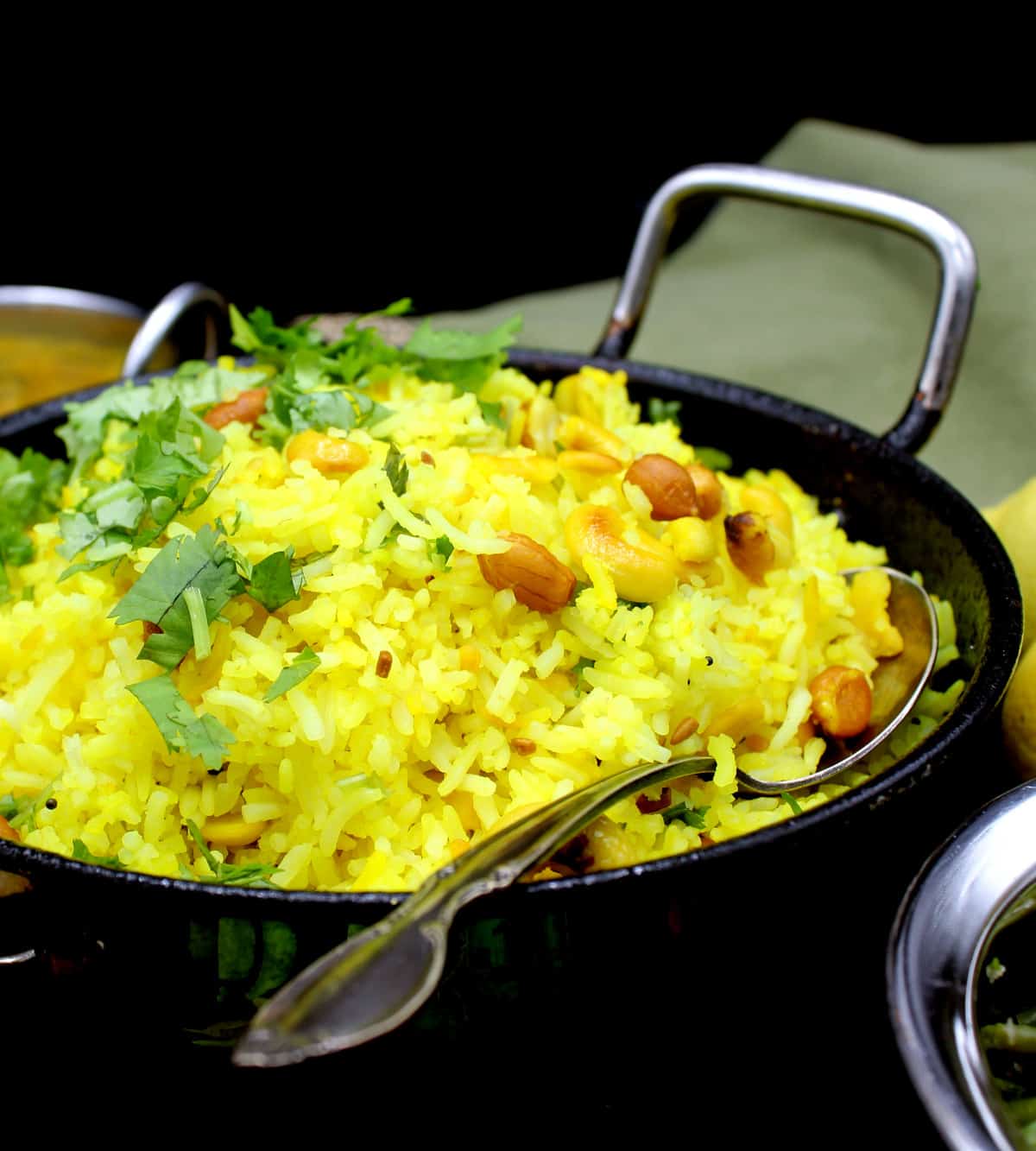 Front partial shot of a steel kadhai with bright yellow lemon rice with mustard seeds, cilantro, peanuts, cashews and red pepper, surrounded by other dishes. Also in the shot is a green and gold napkin and lemons on a black background.